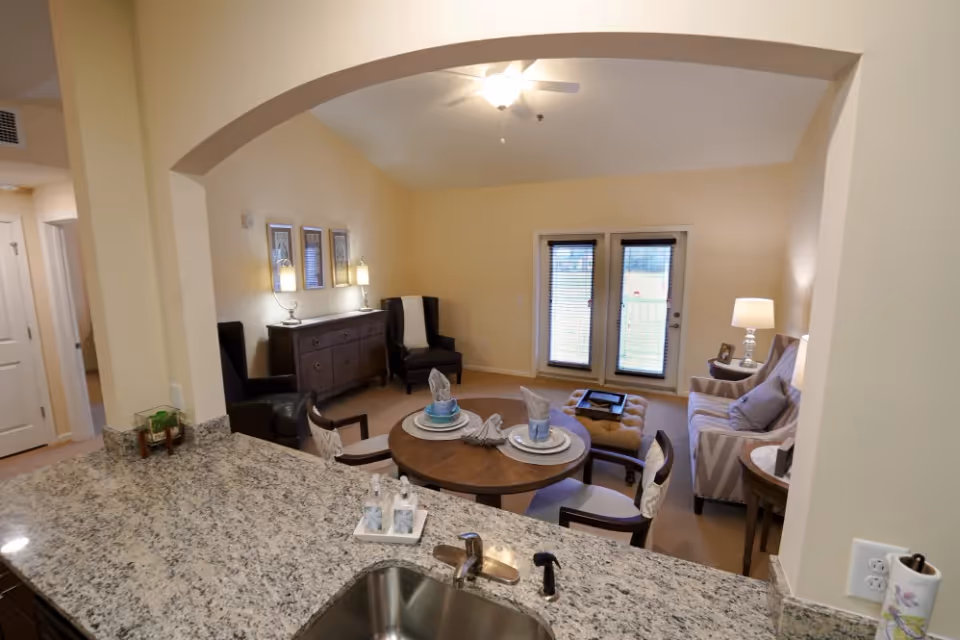 View from a kitchen counter with granite countertop into a living room area featuring a round dining table set for two, two armchairs, a sofa, an ottoman, and a sideboard with lamps and framed pictures on the wall. French doors with blinds lead outside.