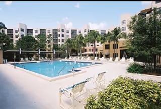 Outdoor swimming pool area surrounded by lounge chairs and palm trees, with a multi-story residential building in the background under a partly cloudy sky.
