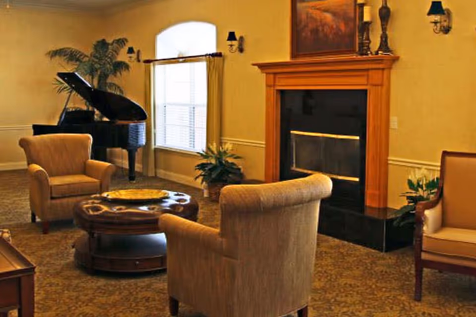 A cozy living room with beige armchairs arranged around a round wooden coffee table. There is a grand piano in the corner near a window with curtains, and a wooden fireplace with decorative items on the mantel. The room has warm lighting and carpeted flooring.
