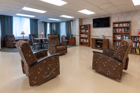 A spacious common area with four brown patterned recliners arranged in a circle on a beige floor. In the background, there are bookshelves filled with books, a flat-screen TV mounted on the wall, a wooden desk with a computer, and a table with chairs near large windows covered with curtains. The room is well-lit with ceiling lights.