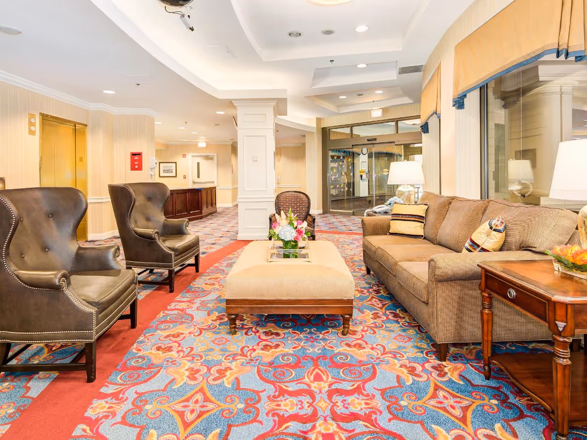Bright furnished lobby with sofas and armchairs arranged around a central ottoman topped with flowers, and a colorful patterned carpet.