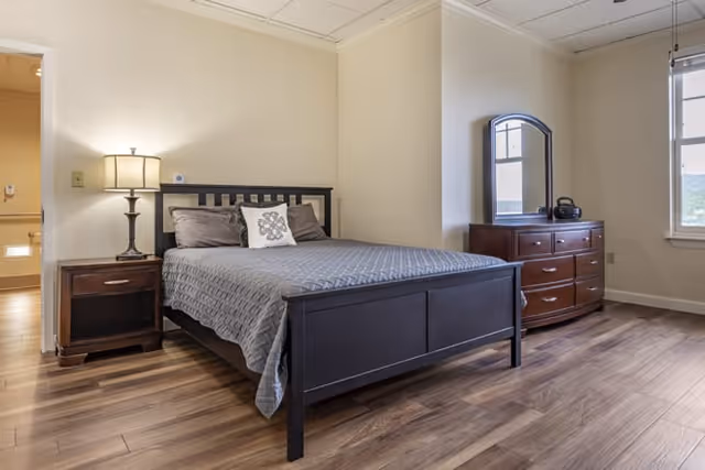 A bedroom with a dark wooden bed frame and gray bedding, a nightstand with a lamp on the left side of the bed, a wooden dresser with a mirror on the right side near a window, and wood flooring throughout the room.