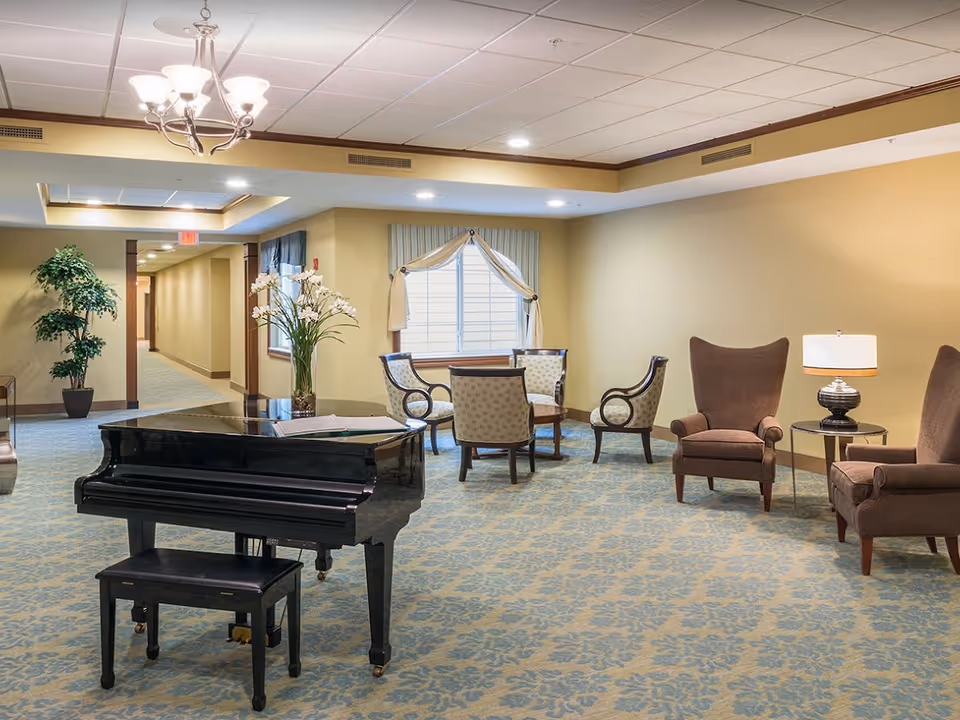 A spacious common area with a black grand piano and matching bench in the foreground. Behind the piano, there is a seating area with four upholstered chairs arranged around a small wooden table near a window with draped curtains. To the right, there are two brown wingback chairs with a round side table and a lamp between them. The room has beige walls, a patterned carpet, recessed lighting, and a chandelier hanging from the ceiling.