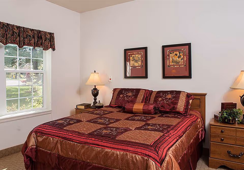 A bedroom in a senior living facility featuring a large bed with a red and brown patterned quilt and matching pillows. There are two framed artworks hanging on the white wall above the bed. On each side of the bed are wooden nightstands with table lamps, a book, and a small plant. A window with a decorative valance allows natural light into the room.