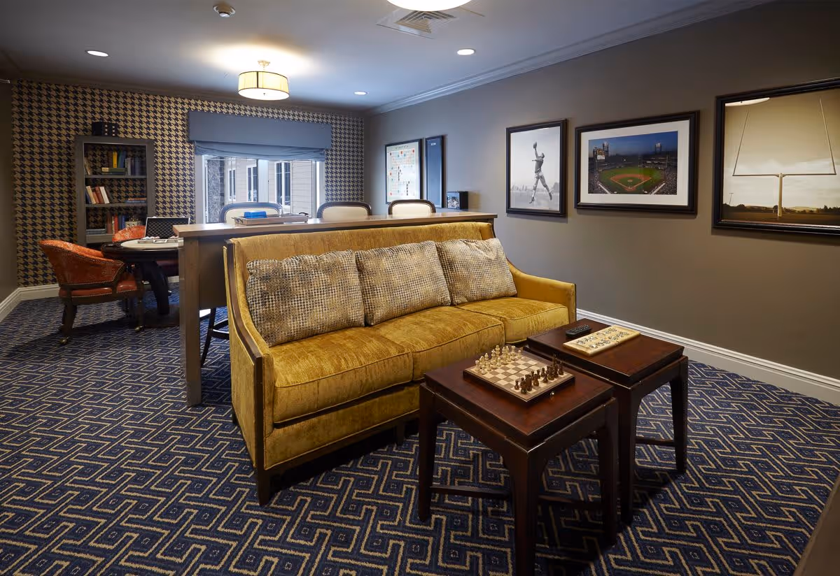 A cozy living room area with a mustard yellow sofa adorned with patterned cushions, two wooden nesting tables with a chessboard and a game board on top, and a patterned blue carpet. Behind the sofa is a tall table with three chairs. The walls are decorated with framed pictures including a baseball player, a baseball stadium, and a football goalpost. A bookshelf and a round table with chairs are visible in the background.