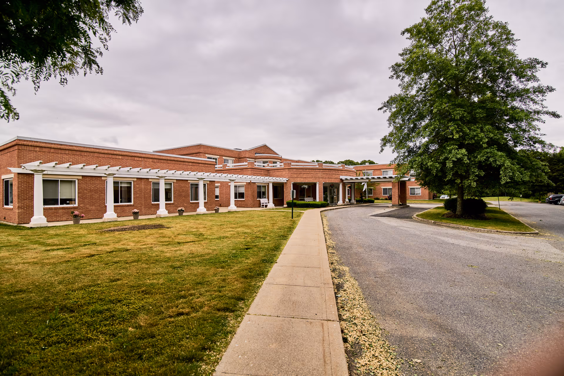 Exterior view of a single-story brick building with white columns and a covered entrance. There is a sidewalk leading to the entrance, a grassy lawn with potted plants, and a large tree on the right side near a curved driveway under a cloudy sky.