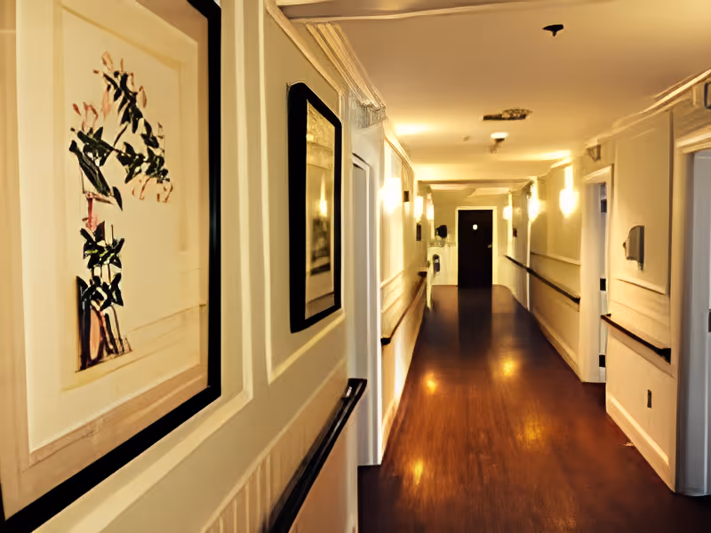 A well-lit hallway in an assisted living facility with wooden flooring, handrails on both sides, framed artwork on the walls, and several doors leading to rooms.