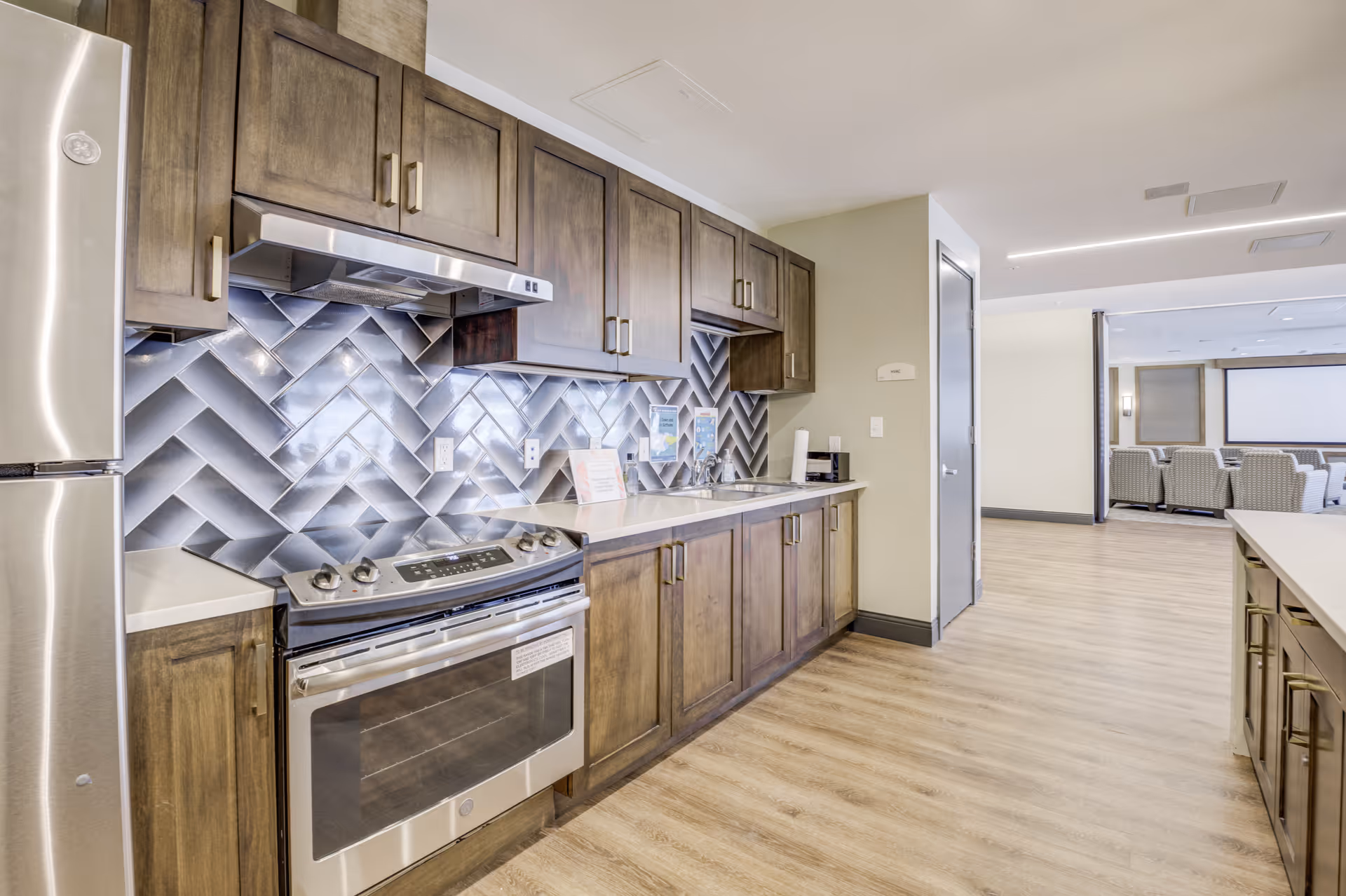 Modern kitchen area with stainless steel refrigerator and stove, wooden cabinets, and a unique herringbone-patterned metallic backsplash. The kitchen opens into a room with chairs and a large screen, suggesting a communal or multipurpose space.