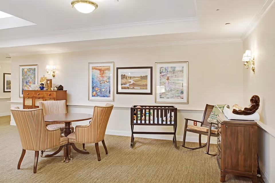 A cozy interior room featuring a round wooden table with four striped upholstered chairs, a wooden crib, a wooden rocking chair with a green blanket, and a wooden dresser with a stuffed animal on top. The walls are decorated with three framed paintings and two wall-mounted light fixtures.