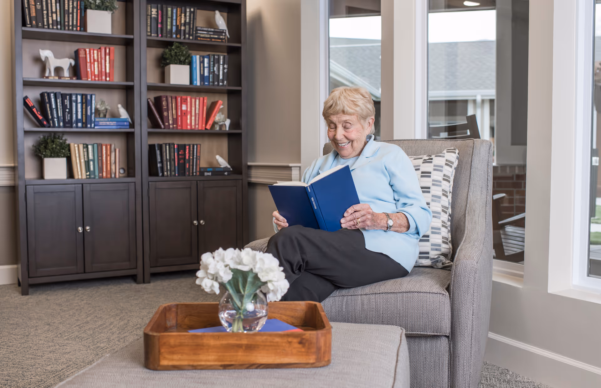 An elderly woman sitting comfortably in a gray armchair in a well-lit room, reading a blue book and smiling. Behind her is a large dark wooden bookshelf filled with books and decorative items. A wooden tray with a glass vase of white flowers is on a nearby ottoman. Large windows let in natural light.