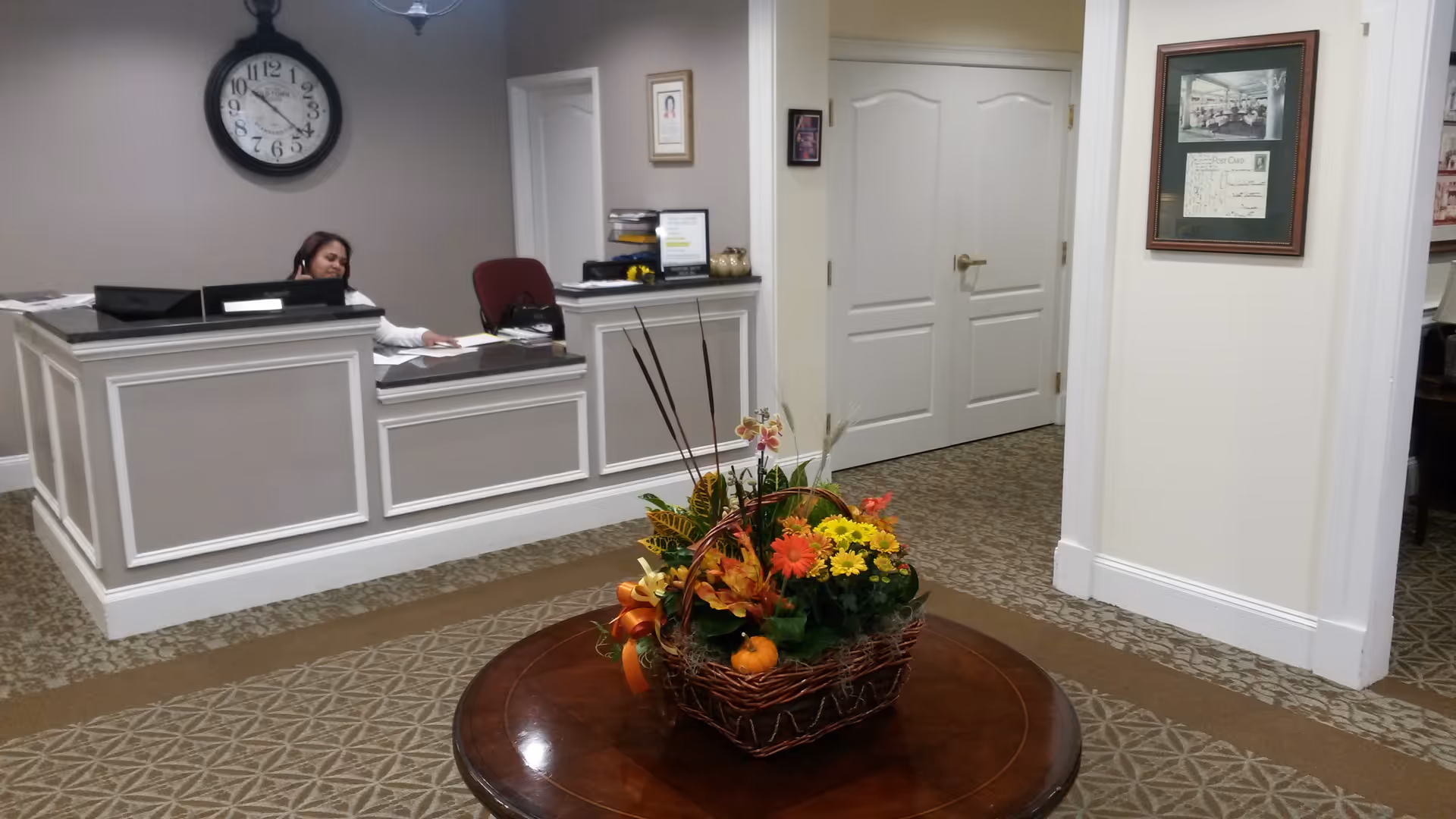Reception area of an assisted living facility with a woman sitting behind a white and gray reception desk. A large round clock is mounted on the wall above the desk. In the foreground, there is a round wooden table with a decorative basket of colorful flowers. The floor is carpeted with a patterned design, and there are white double doors and framed pictures on the walls.