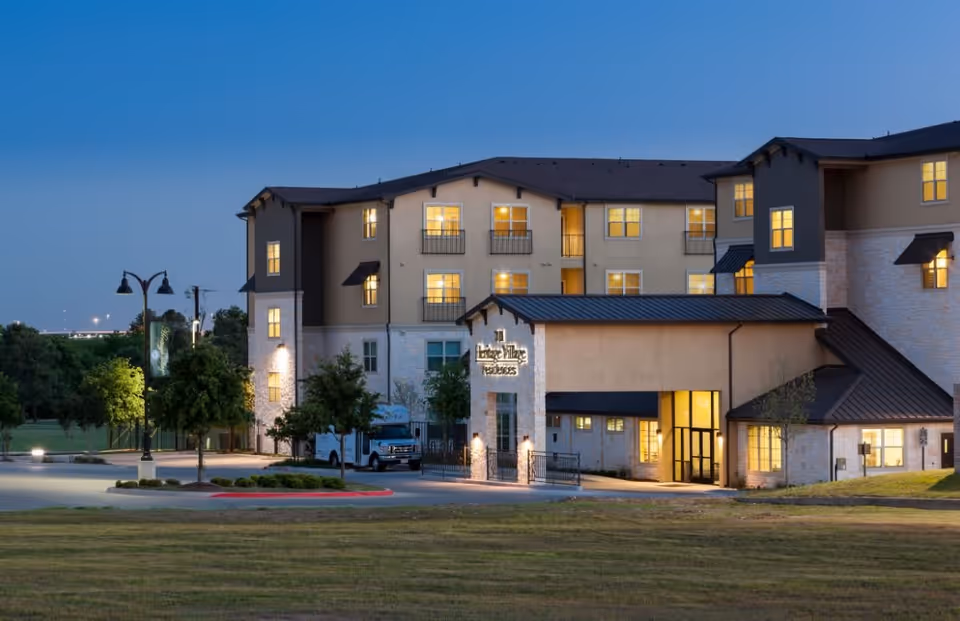 Exterior view of Heritage Village Residences building at dusk with lights on inside, surrounded by trees and a grassy area in the foreground.