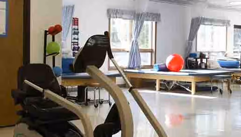 A bright physical therapy or exercise room with large windows covered by light curtains. The room contains exercise equipment including a recumbent bike, therapy tables with exercise balls, and various fitness accessories organized on racks.