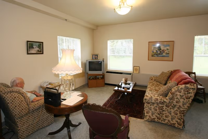 A cozy living room with an elderly person sitting in an armchair. The room features a floral patterned sofa, a wooden coffee table on a dark red rug, a small TV on a wooden stand, two windows with blinds, framed pictures on the walls, and a table with a decorative lamp and some papers.