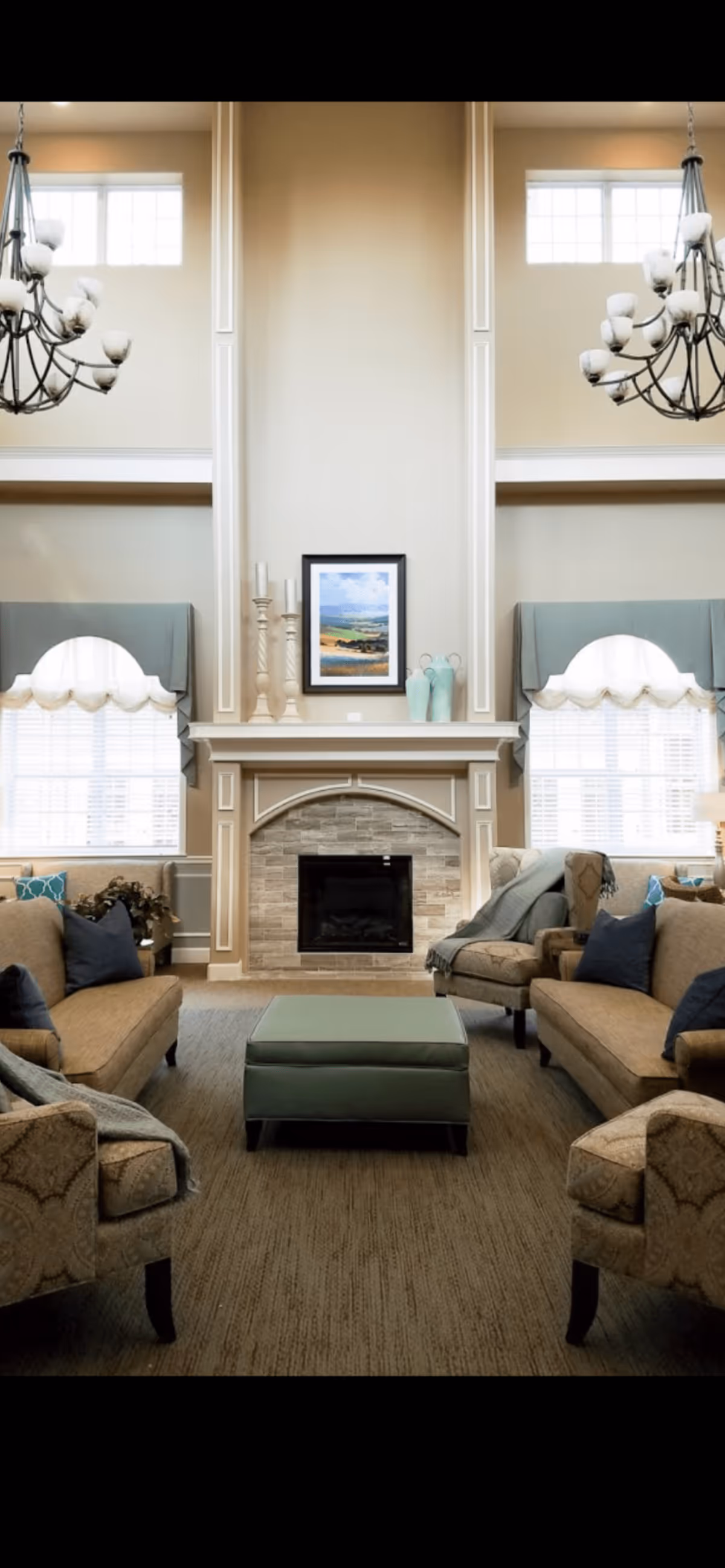 Spacious, elegant common living room with high ceilings, a central fireplace, sofas arranged around an ottoman, and hanging chandeliers.
