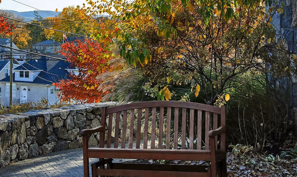 A wooden bench on a stone-paved patio surrounded by autumn foliage including red, yellow, and green leaves. A stone wall borders the patio, and houses and hills are visible in the background under a clear sky.