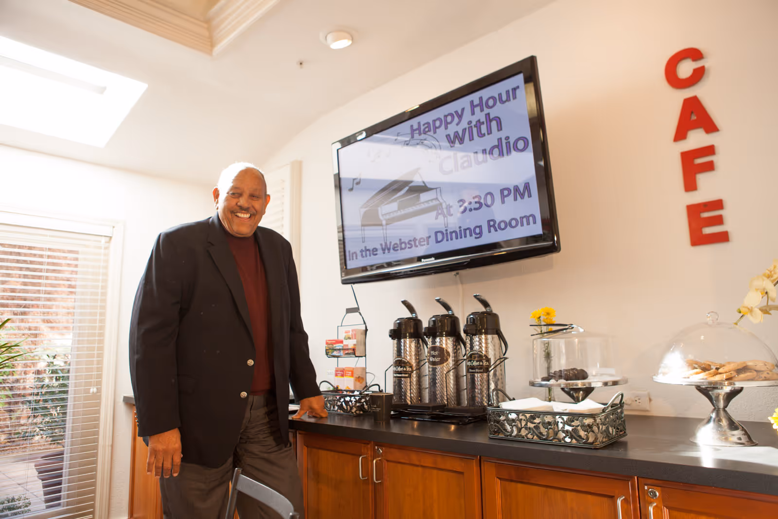 A smiling man stands in a cafe area with a television screen displaying an event announcement.