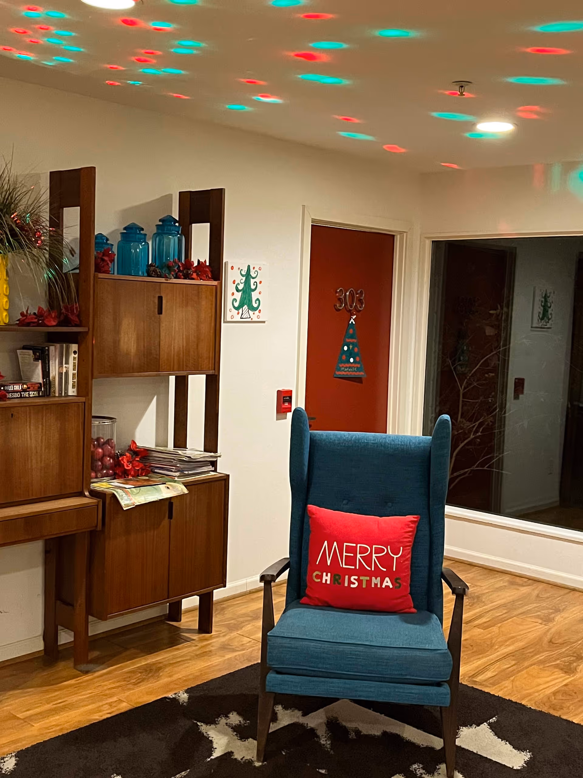 A cozy interior corner of a senior living facility decorated for Christmas. There is a teal armchair with a red pillow that says 'Merry Christmas.' Behind the chair is a wooden shelving unit with blue jars, books, and holiday decorations. The door in the background is red with the number 303 and a Christmas tree decoration. Colorful red and green lights are projected on the ceiling.