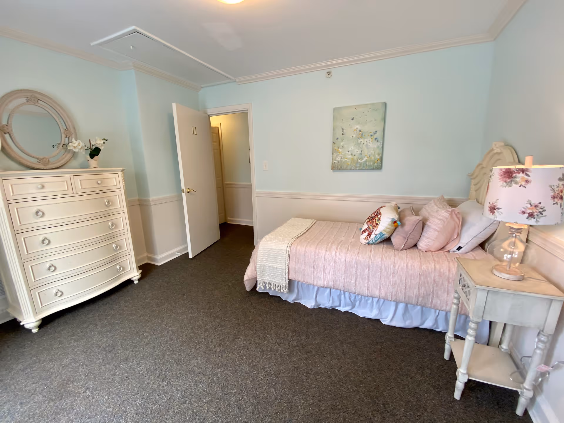 A cozy bedroom in a memory care home featuring a single bed with pink bedding and multiple pillows, a small bedside table with a floral lamp, a white dresser with a round mirror and a vase with flowers, light blue walls with white wainscoting, and an open door numbered 11 leading to a hallway.