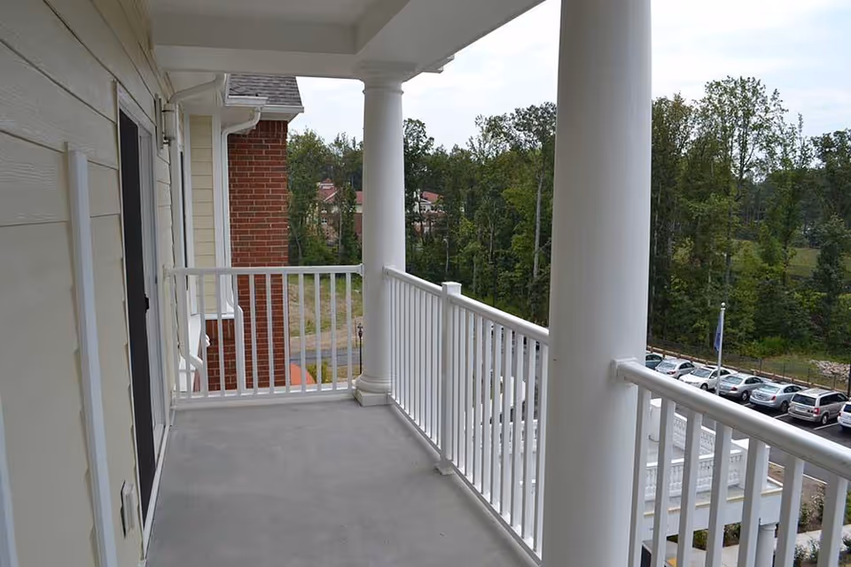 Covered balcony with white railings and columns overlooking a parking lot and trees.