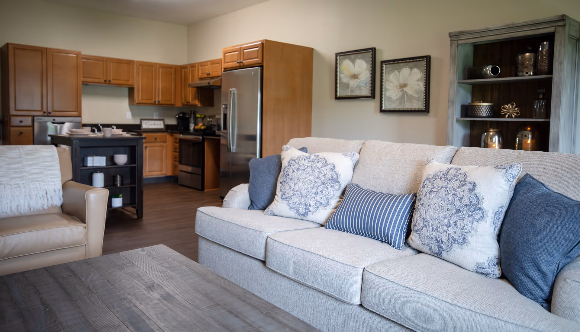A cozy living room area with a beige sofa adorned with decorative pillows and a beige armchair with a white throw blanket. In the background, there is a kitchen with wooden cabinets, a stainless steel refrigerator, stove, and a small black kitchen island. On the wall above the sofa, there are two framed floral artworks, and to the right, a wooden shelving unit with decorative items.