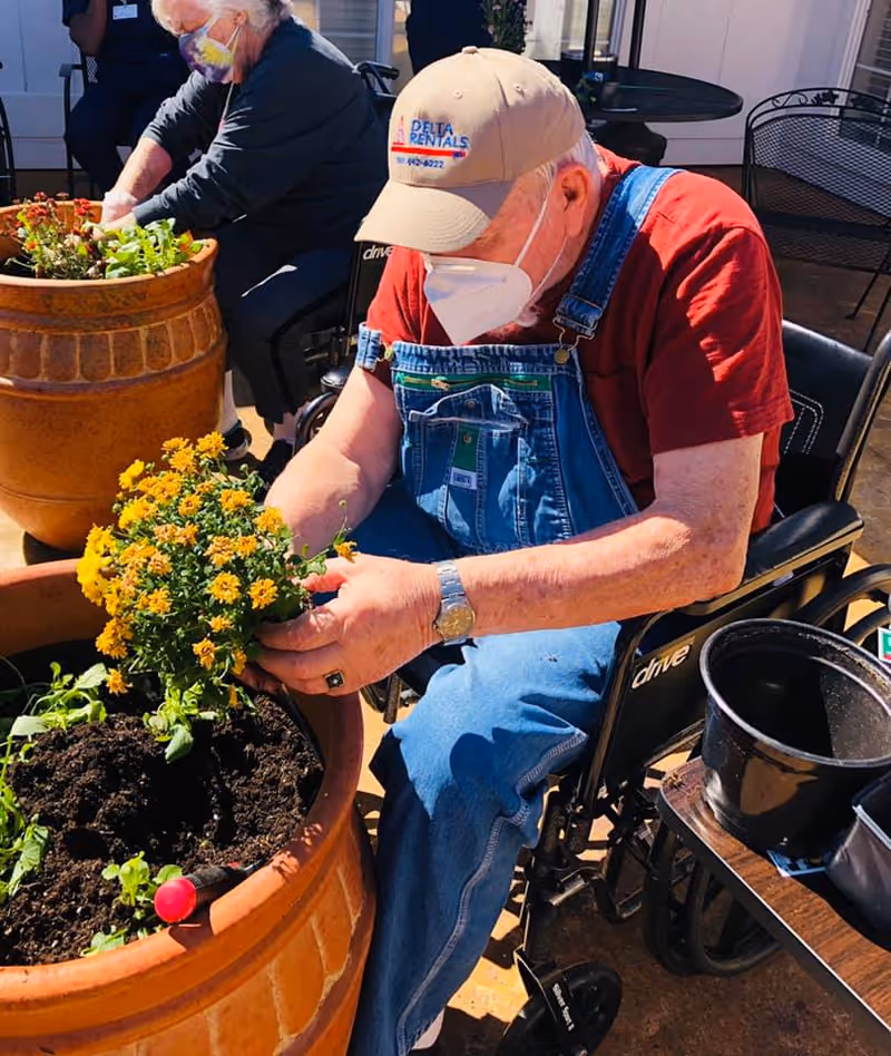 An elderly man wearing a beige cap, red shirt, blue overalls, and a white face mask is sitting in a wheelchair while planting yellow flowers in a large terracotta pot outdoors. Another elderly person, also wearing a face mask, is visible in the background tending to plants in a similar pot. There are gardening tools and pots nearby on a table.