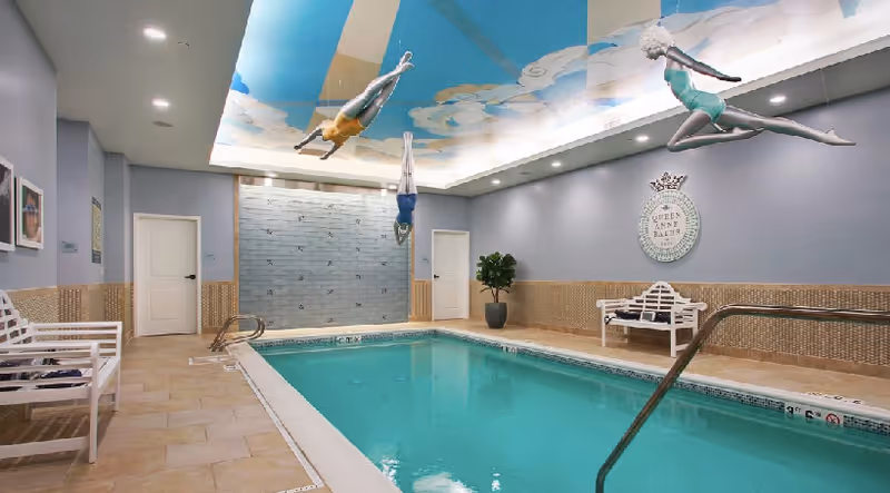 Indoor swimming pool area with a rectangular pool surrounded by beige tiled flooring and light purple walls. There are white benches on either side of the pool, a potted plant, and a decorative wall plaque that reads 'Queen Anne Rodgers Park'. The ceiling features a mural of a blue sky with clouds and three sculptures of divers in mid-air positioned above the pool.