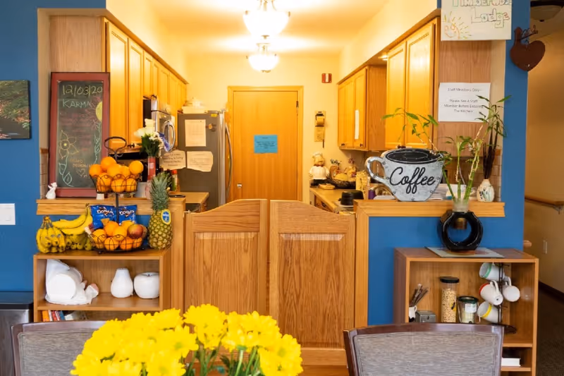 A bright communal kitchen and serving area with wooden cabinets, fruit bowls and a 'Coffee' sign, seen over a pass-through counter with a dining table and yellow flowers in the foreground.