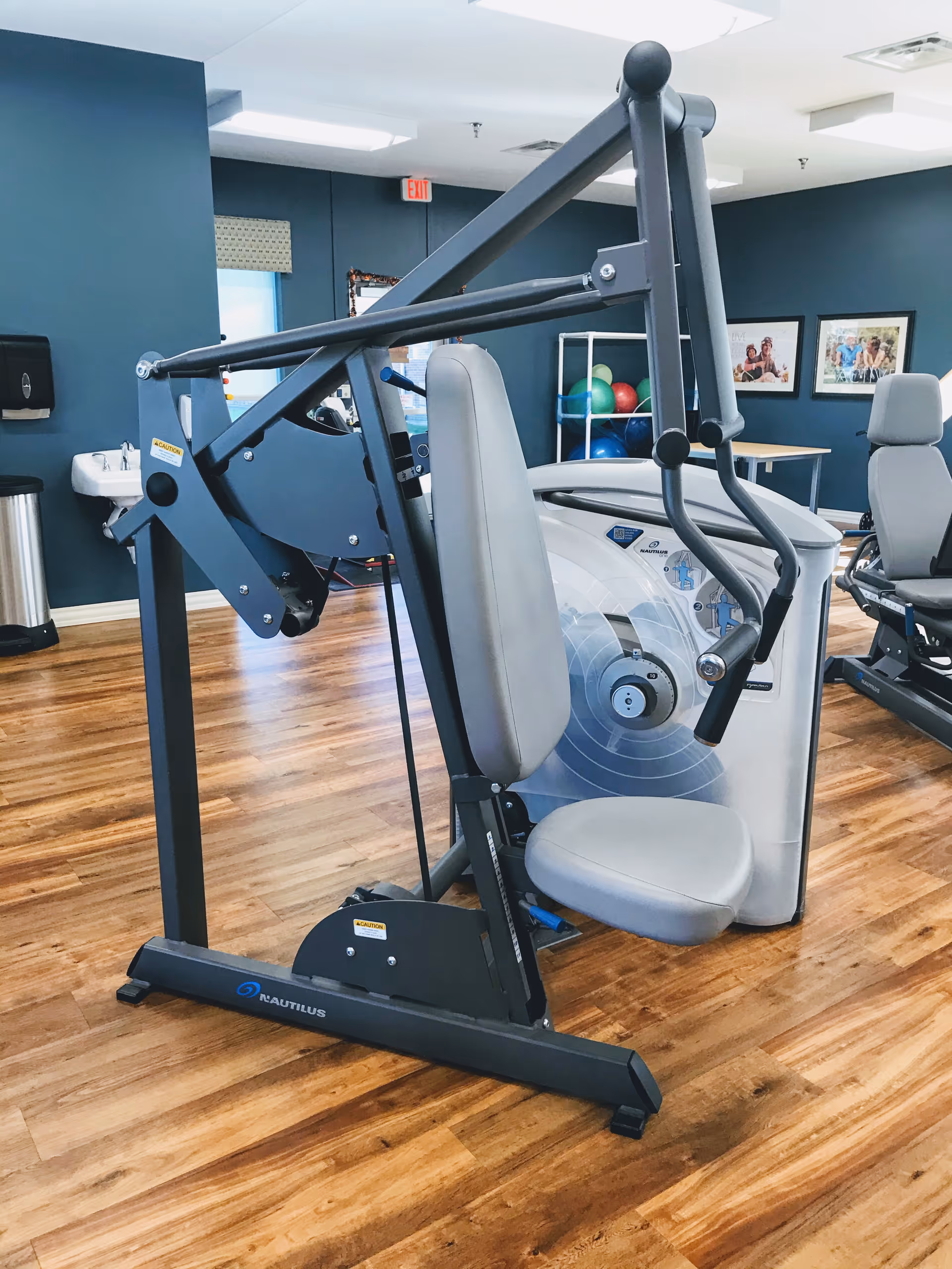 A fitness machine with a padded seat and backrest in a room with wooden flooring and blue walls. In the background, there are exercise balls on a rack, framed pictures on the wall, and another exercise machine.