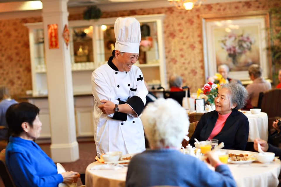 A chef in a white uniform and tall chef hat smiling and interacting with elderly residents seated at a round dining table in a warmly decorated dining room with floral wallpaper and framed artwork.