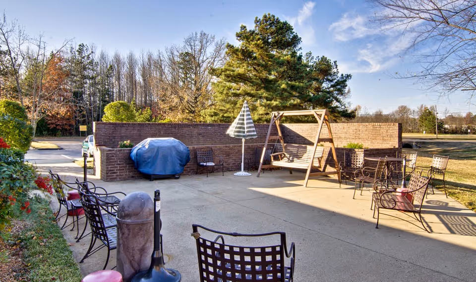Outdoor patio area with metal chairs and benches arranged around a concrete surface. There is a wooden swing, a covered grill, and a striped umbrella. Trees and a brick wall surround the patio, with a clear blue sky overhead.