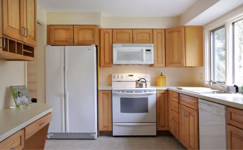 Kitchen with light wood cabinets, white refrigerator, stove with microwave above, dishwasher, and a sink under a large window.