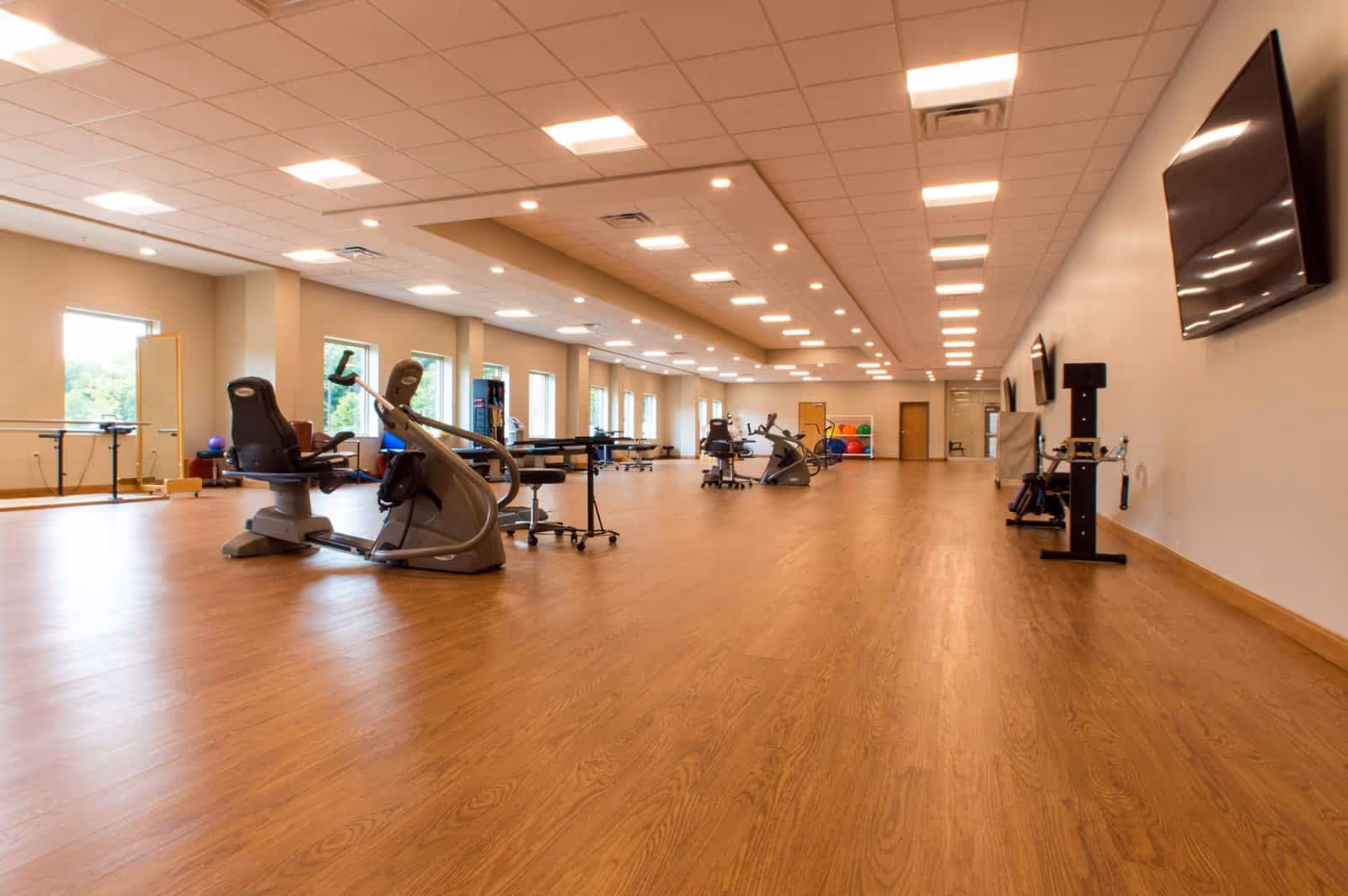 A spacious rehabilitation or physical therapy room with wooden flooring, exercise equipment including stationary bikes, therapy tables, colorful exercise balls, and multiple large windows letting in natural light. The ceiling has recessed lighting, and there are several wall-mounted televisions on the right side.
