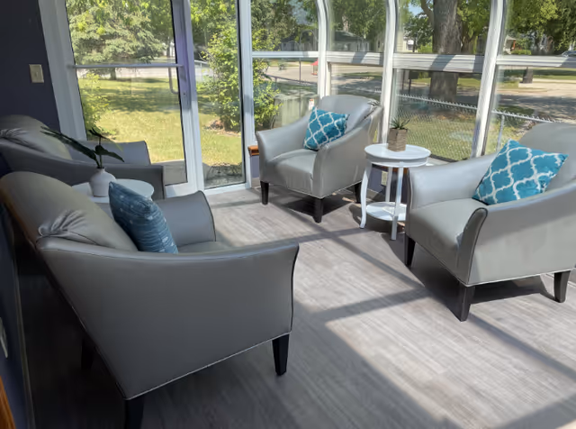 A bright sunroom with four gray armchairs arranged around a small white round table. Each chair has a decorative blue patterned pillow. The room has large windows letting in natural light and a view of green trees and grass outside.