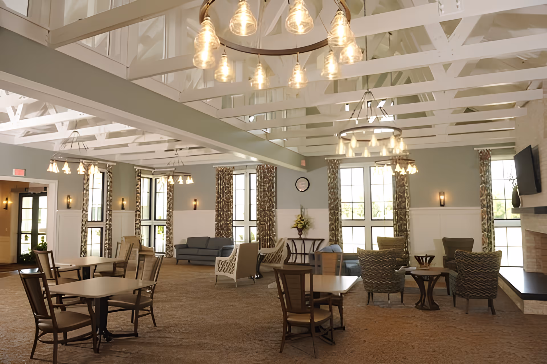 A spacious and well-lit common area with multiple seating arrangements including tables with chairs and comfortable armchairs. The room features large windows with patterned curtains, exposed white ceiling beams, and multiple hanging light fixtures. There is a fireplace with a mounted TV on the right side and a small table with a flower arrangement against the far wall.