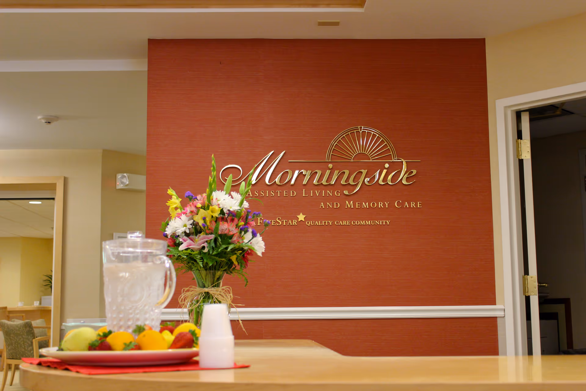 Reception area of Morningside Assisted Living and Memory Care with a red textured wall featuring the facility's gold logo and name. A vase with colorful flowers and a pitcher of water with cups are on the counter in the foreground.