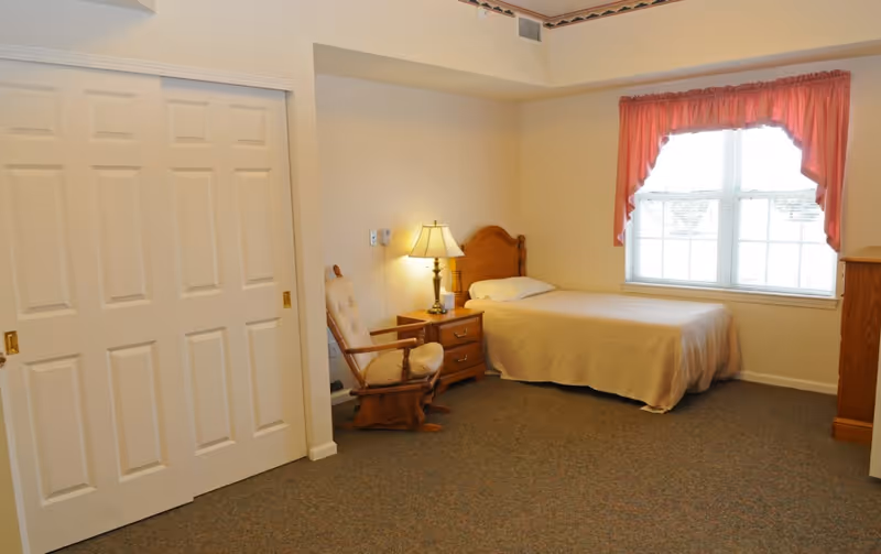 Simple senior living bedroom with a single bed, wooden headboard, nightstand and lamp, a cushioned rocking chair, and a window with a pink valance.