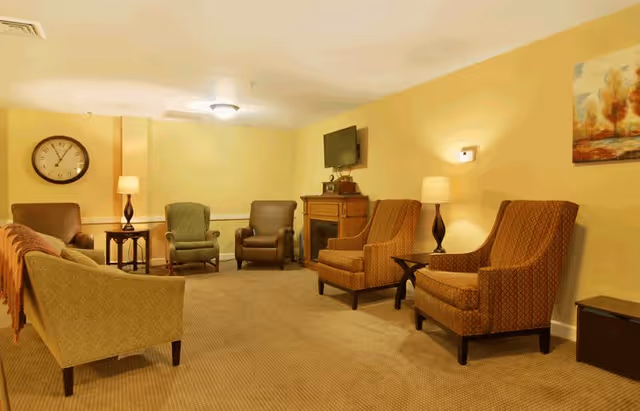 A cozy senior living facility common area with several upholstered armchairs arranged around the room. The walls are painted a warm yellow, and there are two table lamps providing soft lighting. A wall clock is visible on the left wall, and a flat-screen TV is mounted above a wooden fireplace on the far wall. A framed painting hangs on the right wall.