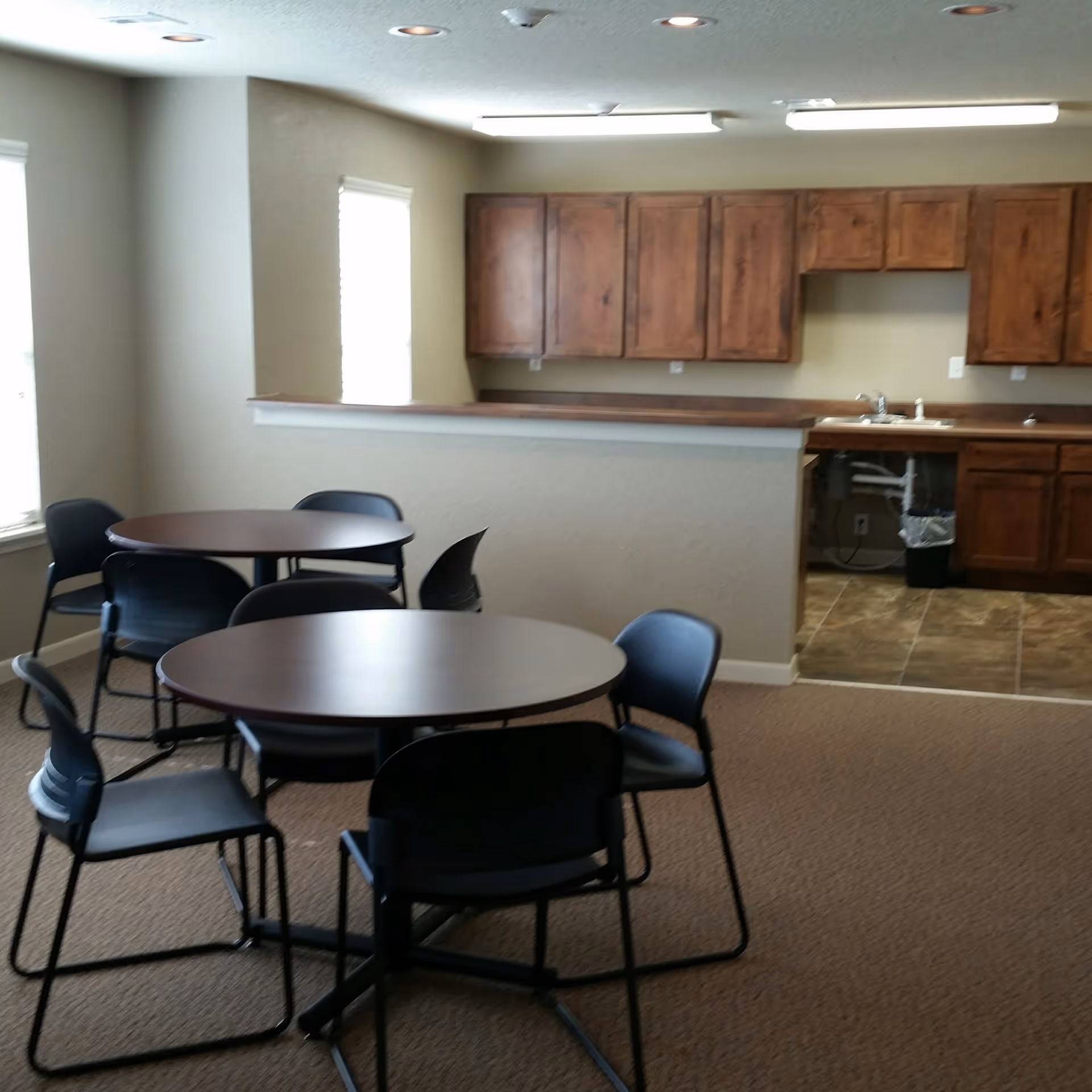 Small communal dining area with round tables and chairs and a kitchenette with wooden cabinets in the background.
