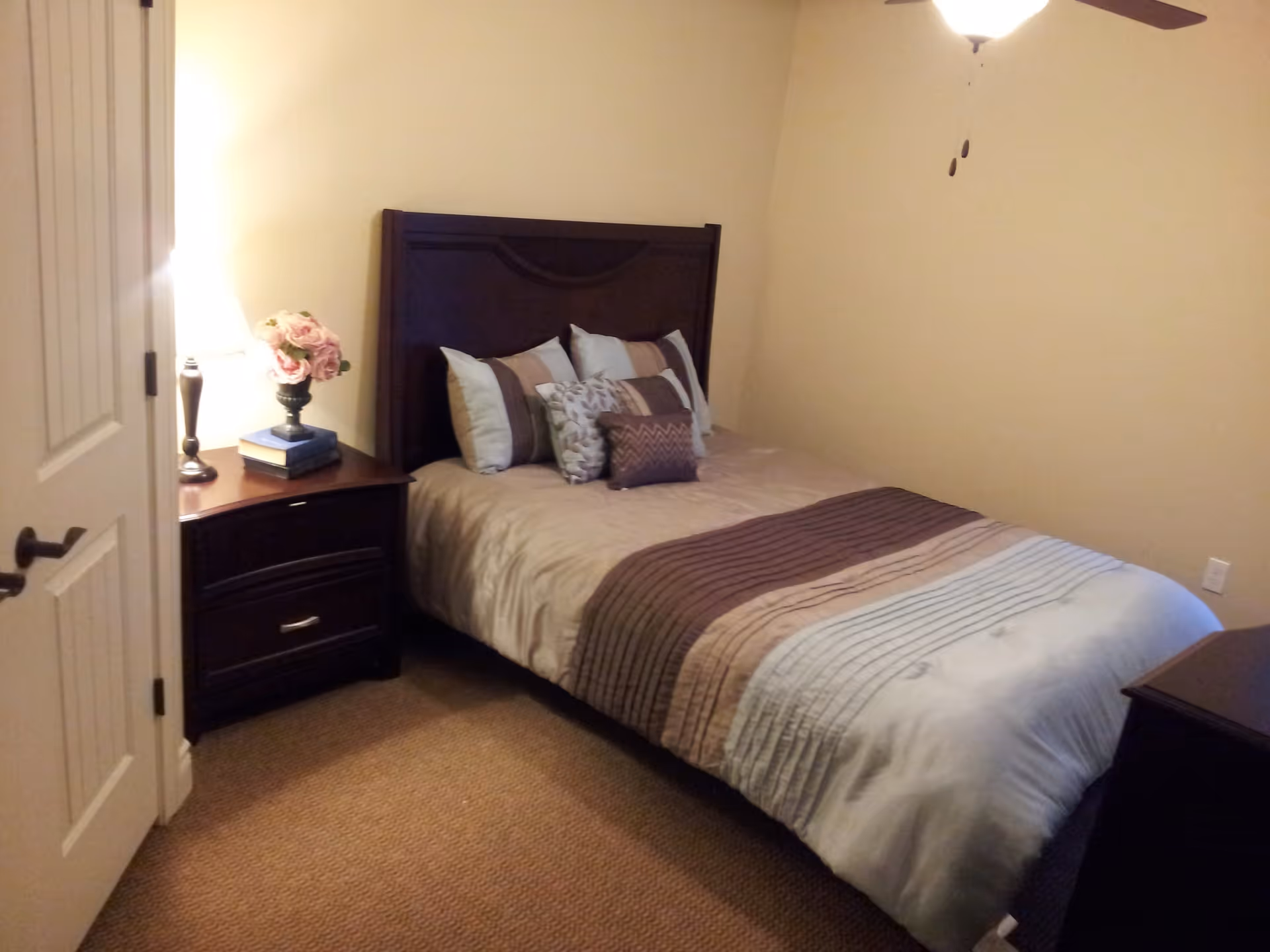 A small bedroom with a neatly made bed featuring a striped comforter and multiple pillows. There is a dark wooden headboard and matching nightstand with a lamp and a vase of flowers on it. The room has beige walls and carpeted floor.