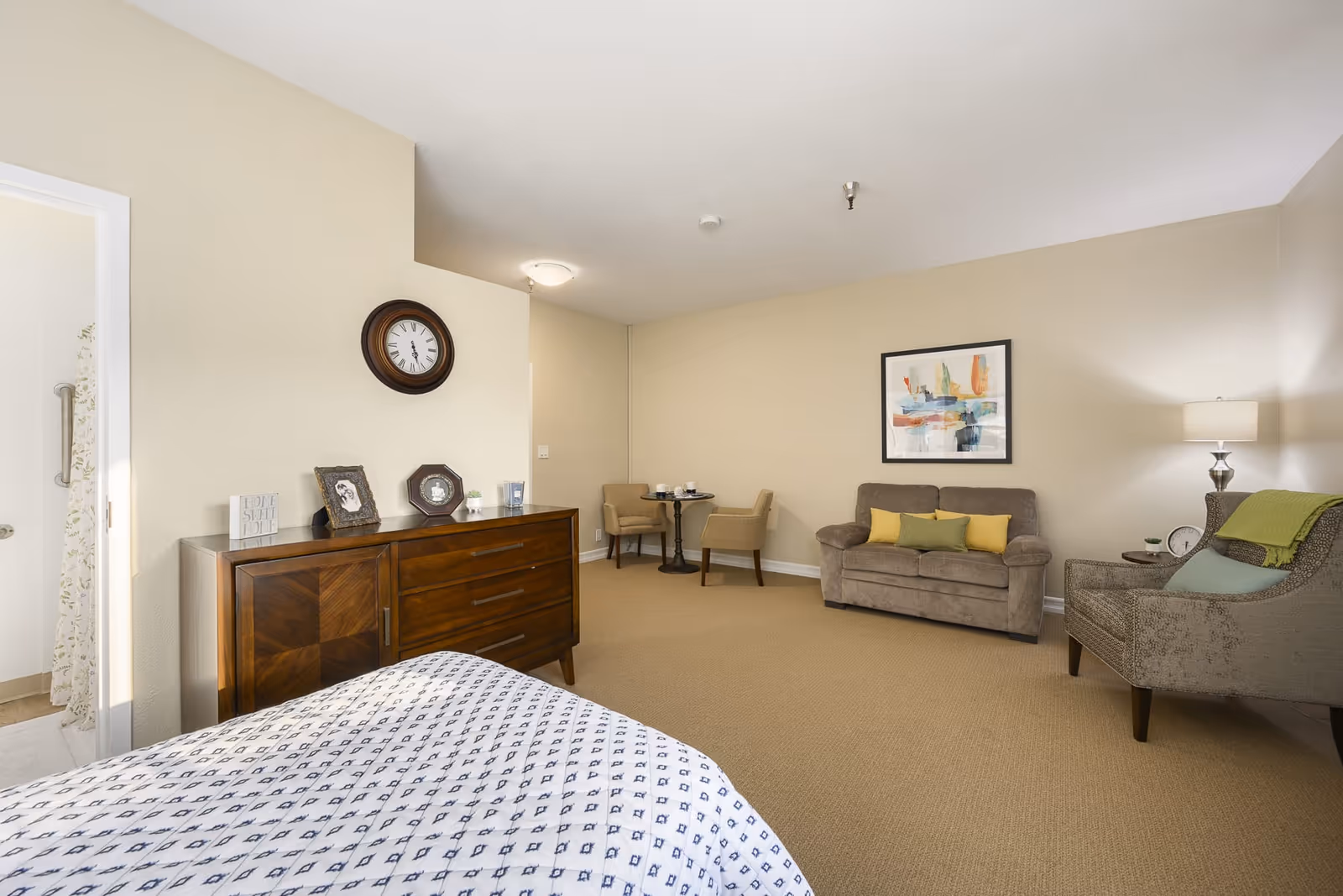 Spacious senior living bedroom with a bed in the foreground, wooden dresser, and a seating area with a sofa and chairs.