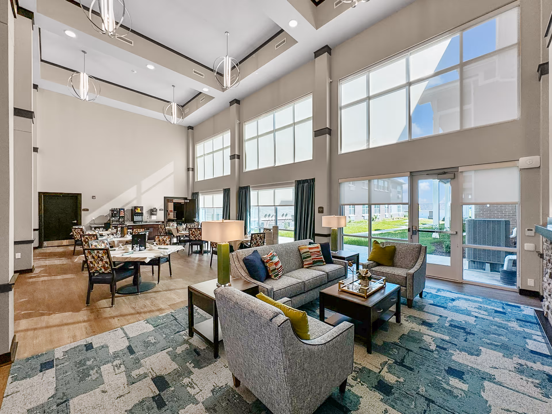 A spacious senior living facility common area with high ceilings and large windows letting in natural light. The room features a seating area with a gray sofa, two armchairs with colorful pillows, a coffee table, and side tables with lamps. In the background, there are dining tables and chairs arranged neatly, and a beverage station against the far wall. The floor has a combination of wood and patterned carpet, and there is a glass door leading outside to a garden area.