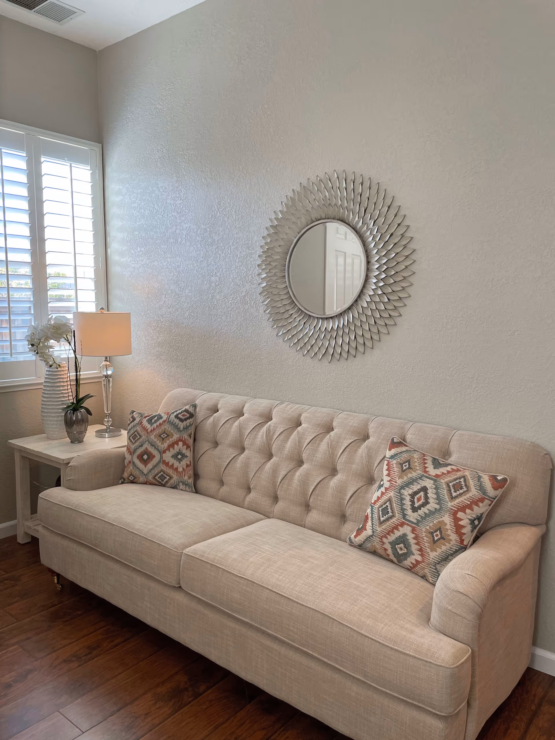 Beige tufted sofa with patterned throw pillows beneath a decorative round mirror, beside a side table with a lamp and window shutters.