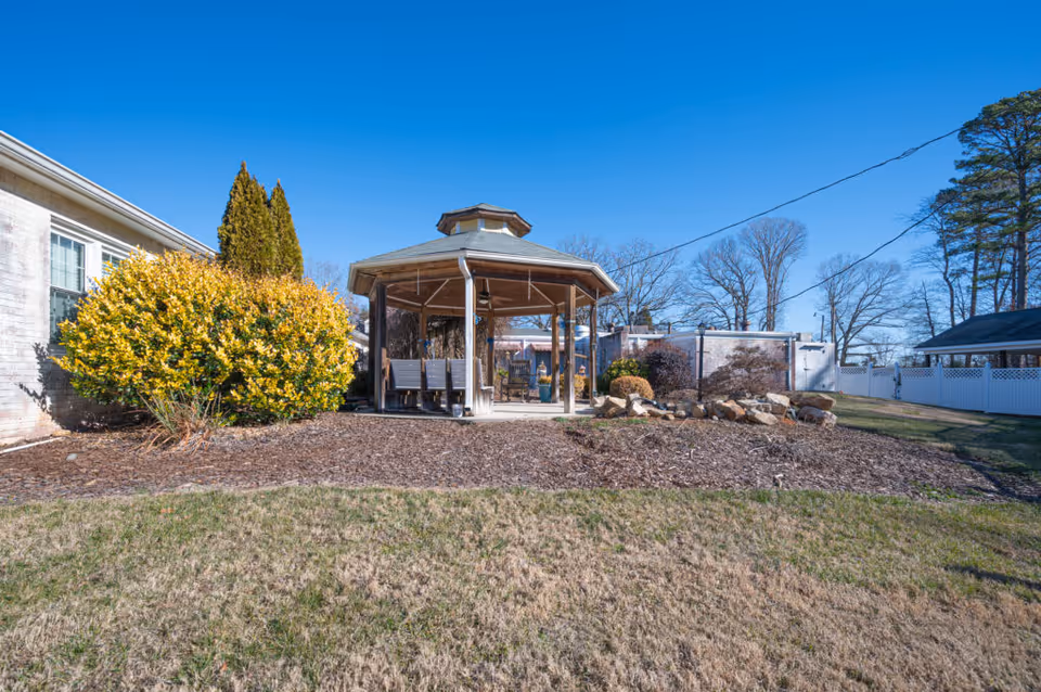 Outdoor view of a senior living facility garden area featuring a wooden gazebo with benches inside, surrounded by bushes, rocks, and a grassy lawn under a clear blue sky.