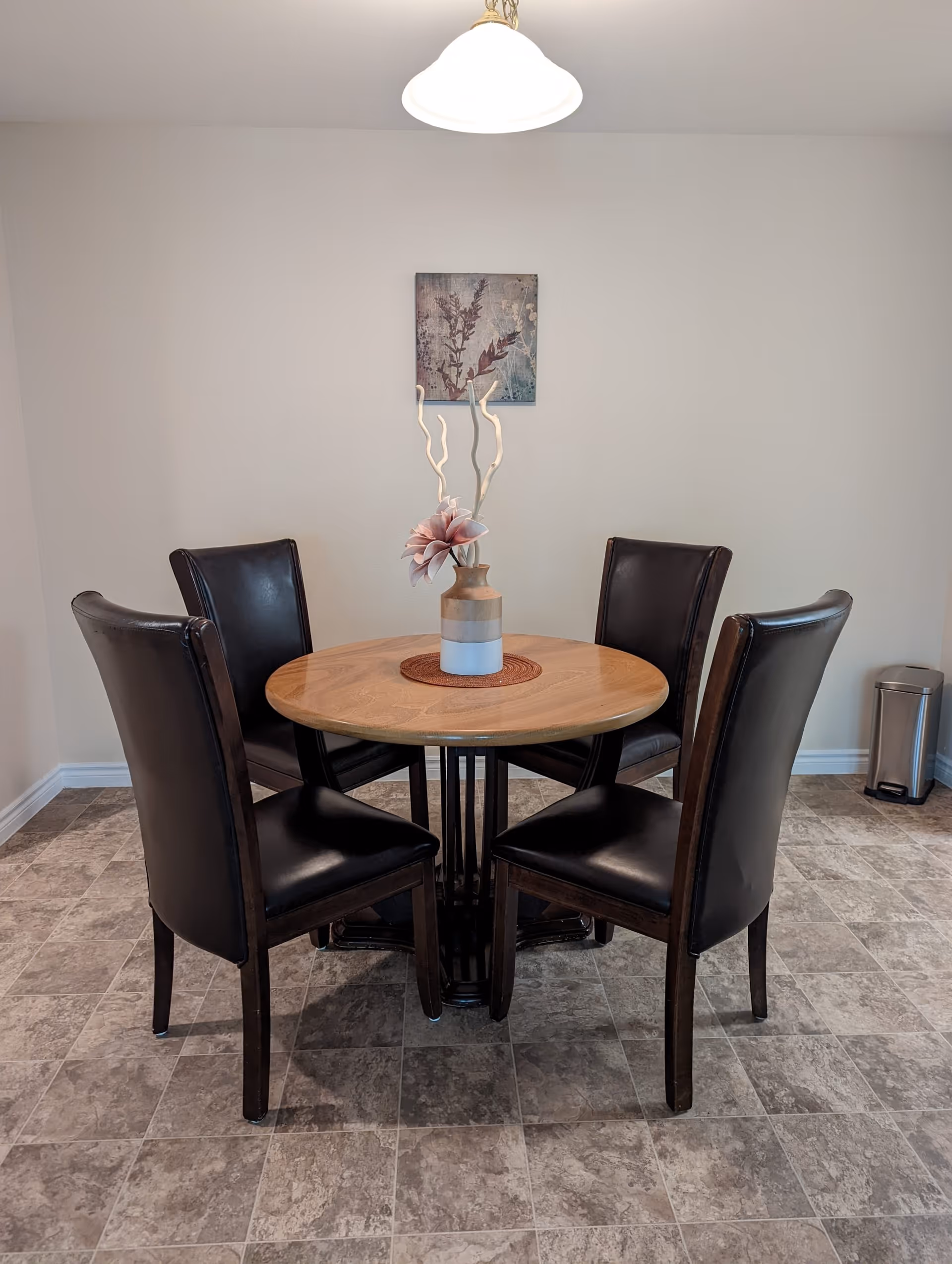 A small round wooden dining table with a vase containing decorative flowers and branches in the center. Four dark brown leather chairs surround the table. The room has tiled flooring, a light fixture hanging from the ceiling, a small framed artwork on the wall, and a stainless steel trash can in the corner.