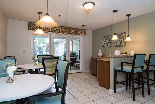 A dining area with round white tables and chairs with teal cushions. There is a small kitchen counter with bar stools and pendant lights hanging above. Sliding glass doors lead to an outdoor patio area. The room has tiled floors and neutral-colored walls.