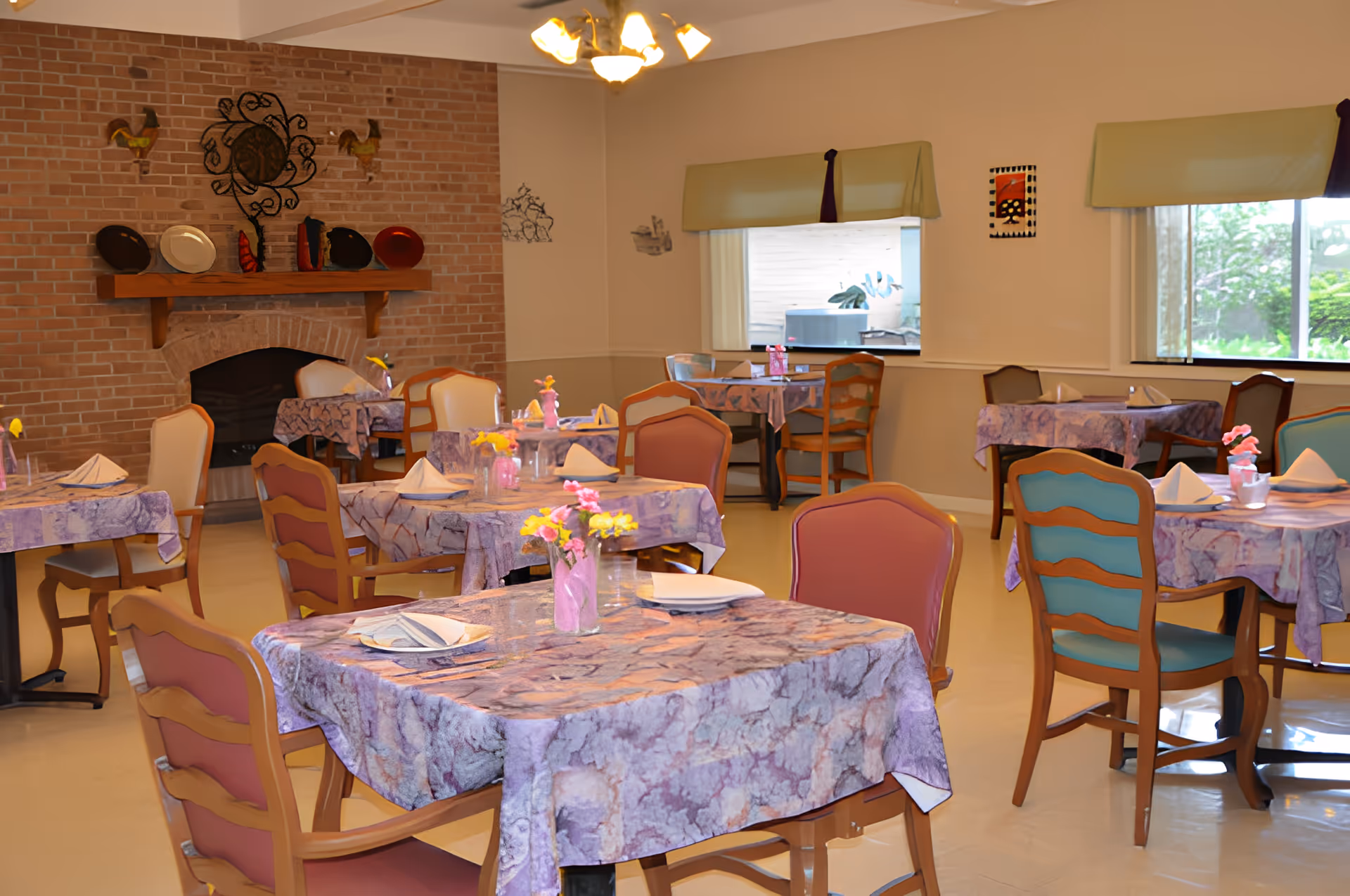 Dining room with multiple tables set with purple tablecloths, floral centerpieces, and wooden chairs in front of a brick fireplace and windows.