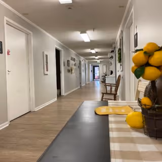 A long, well-lit hallway in a senior living facility with light-colored walls and wood flooring. Several doors line the hallway, and a few chairs are placed along the walls. In the foreground, there is a table with a checkered tablecloth and a decorative arrangement of artificial lemons.