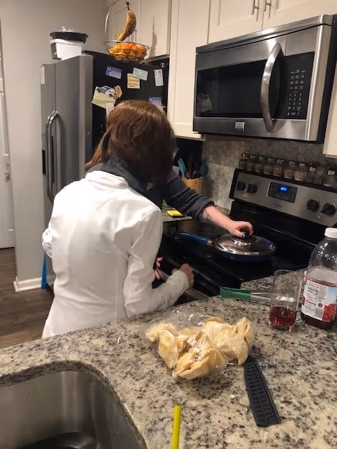 Two people in a kitchen preparing food on a stove. One person is wearing a white jacket and the other is adjusting a pan on the stove. The kitchen has a granite countertop with a bag of chips, a glass with red liquid, and a bottle of juice. Stainless steel appliances including a microwave and refrigerator are visible.