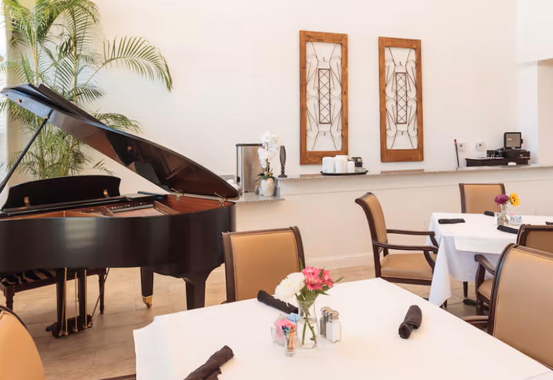 Dining room with a grand piano, set tables with white tablecloths and floral centerpieces, and framed wall decor.